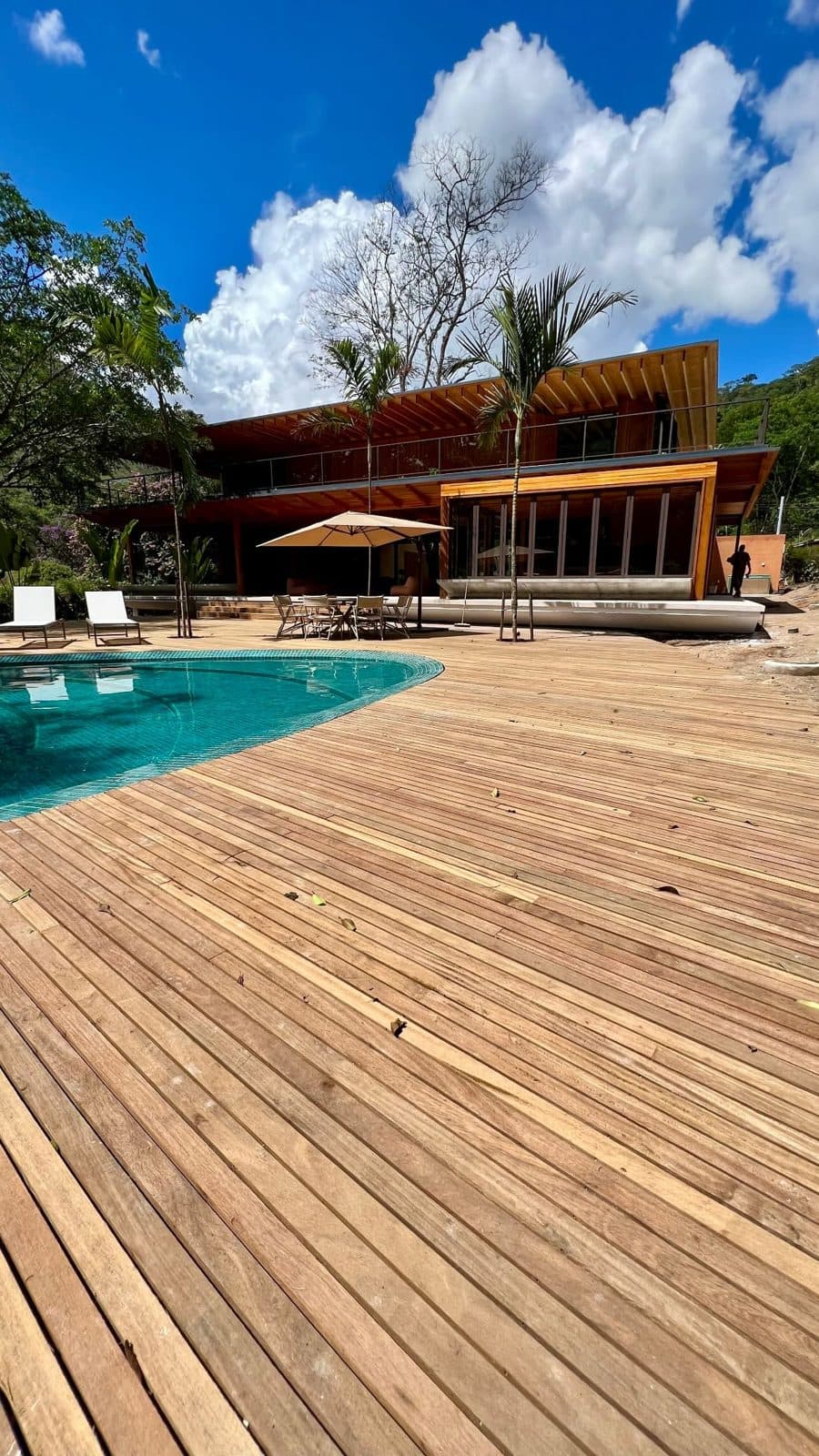 Fachada da casa vista do deck de madeira, com a piscina curva e o céu azul