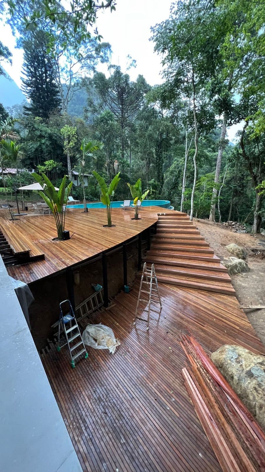 Deck curvo de madeira natural com escadas e canteiros, piscina ao fundo