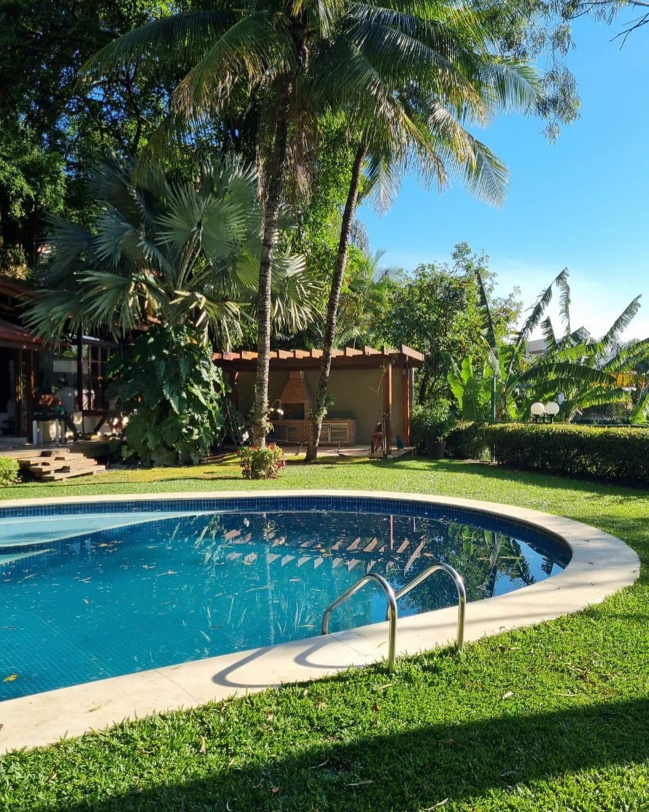Jardim com piscina oval e palmeiras, casa com pergolado ao fundo